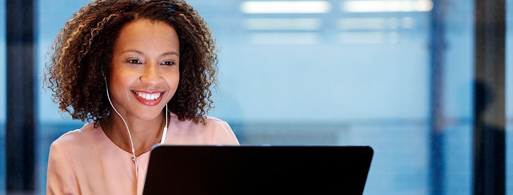 A smiling woman with headphones in an office setting facing a laptop screen.