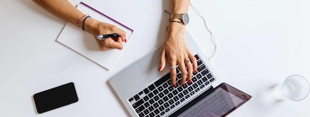 A person taking notes on a piece of paper based on information on a laptop.