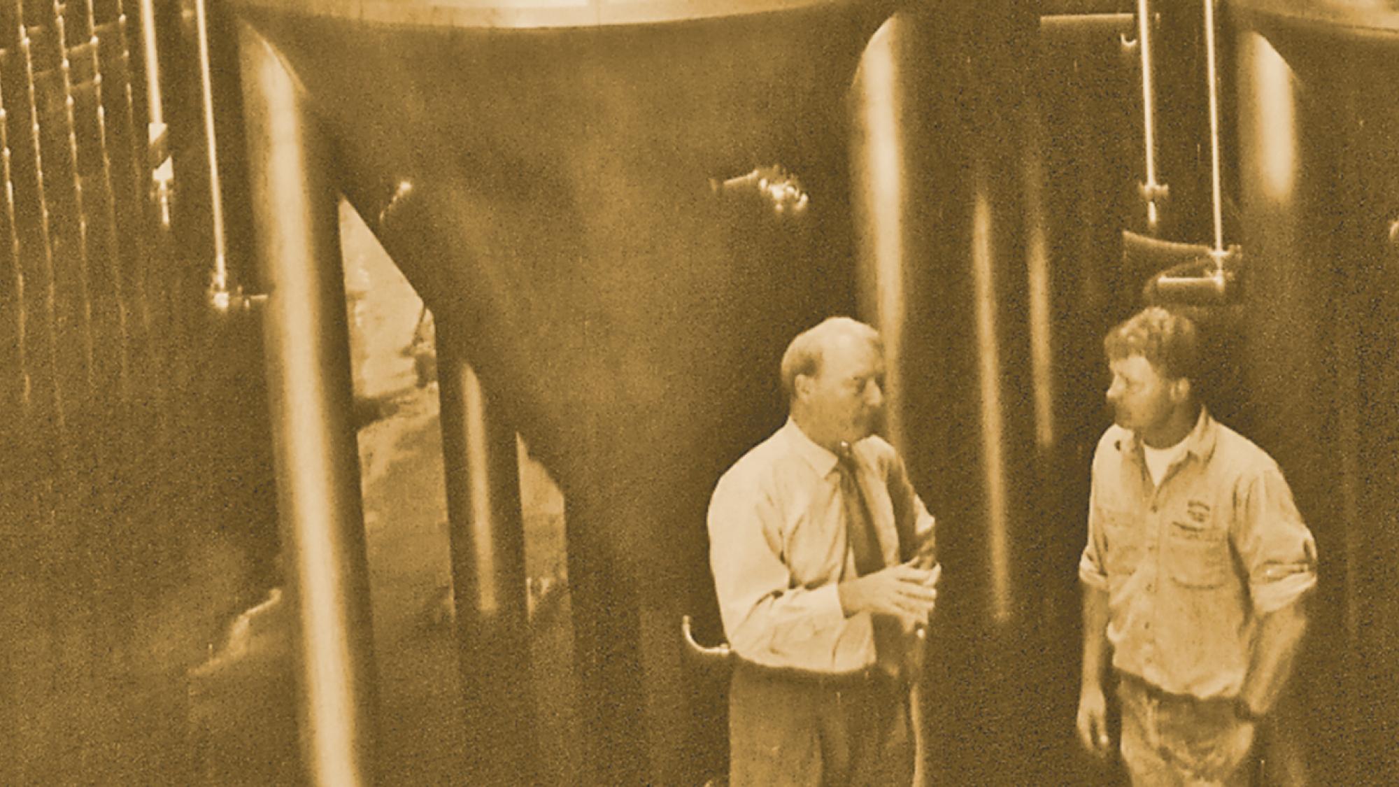 Two men talking beside large brewery tanks, sepia-toned industrial scene