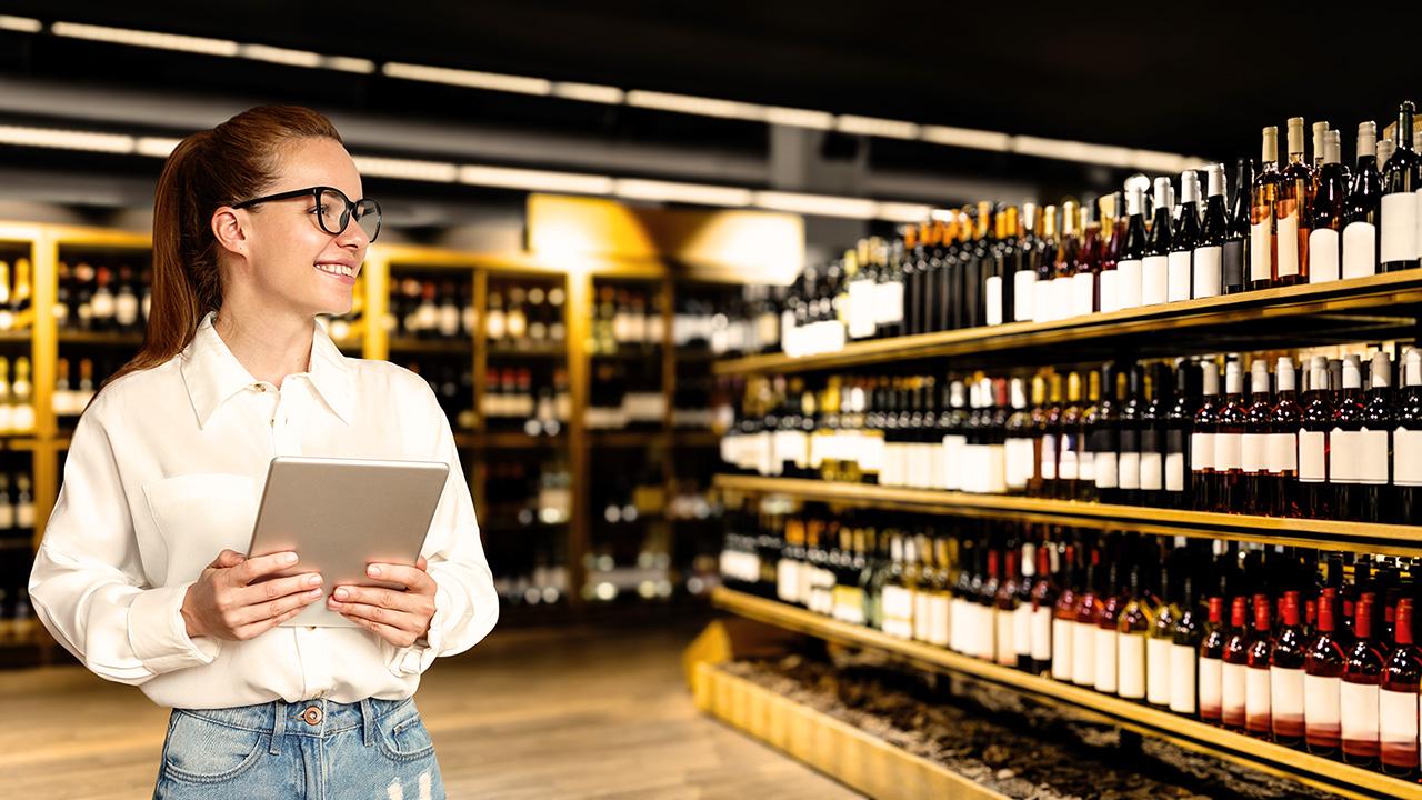 a woman holding a tablet looks at rows of wine bottles in a wine store
