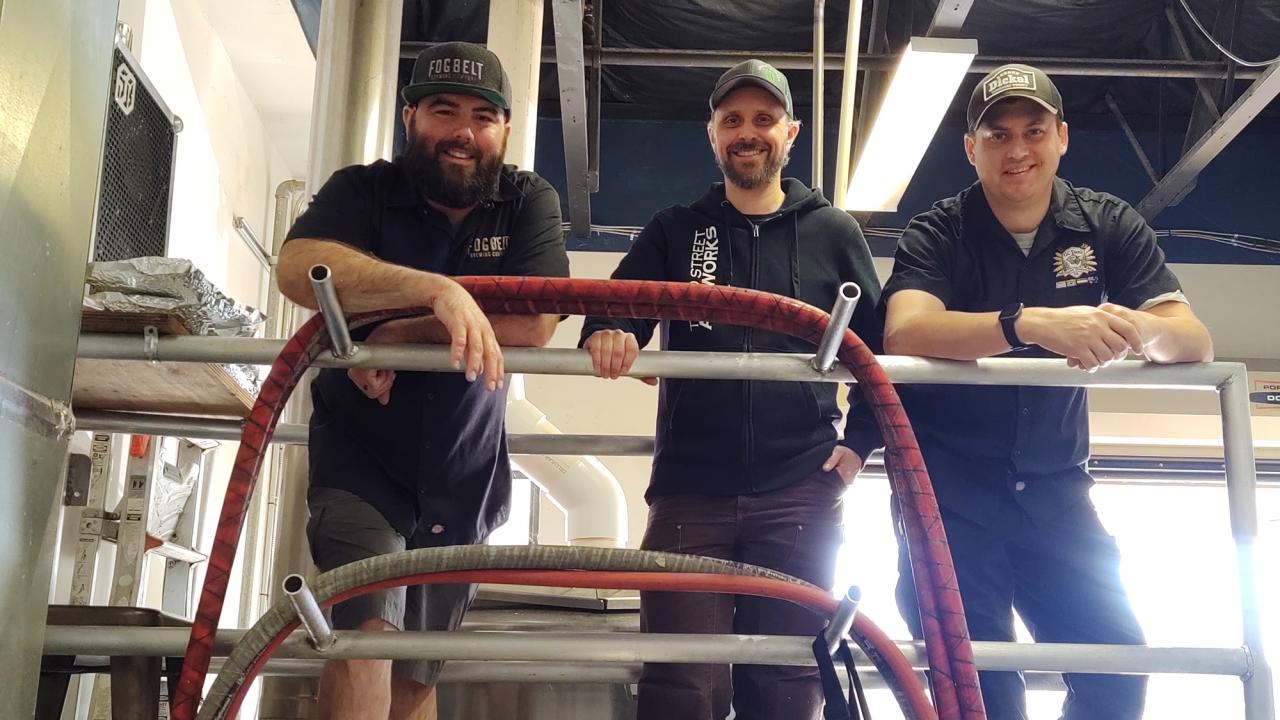Remy Martin, Tyler Laverty and Andy Hooper on the brewdeck at Fogbelt Brewing during the collaboration day. Beer is boiling in the kettle behind them. Remy, Tyler and Andy on the catwalk at the brewery