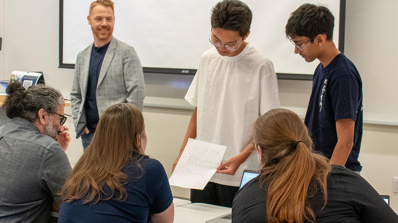 Students present a paper to classmates while a teacher smiles (photo)