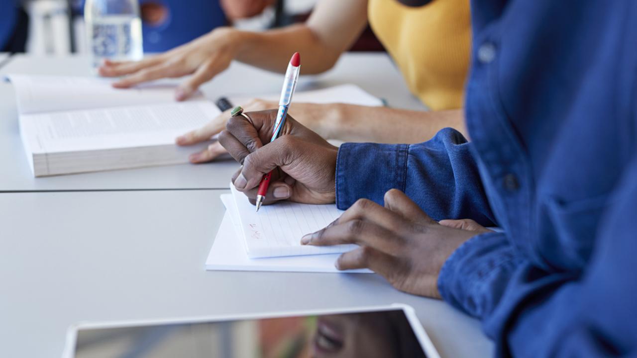 Man writing in book while sitting with friend at desk