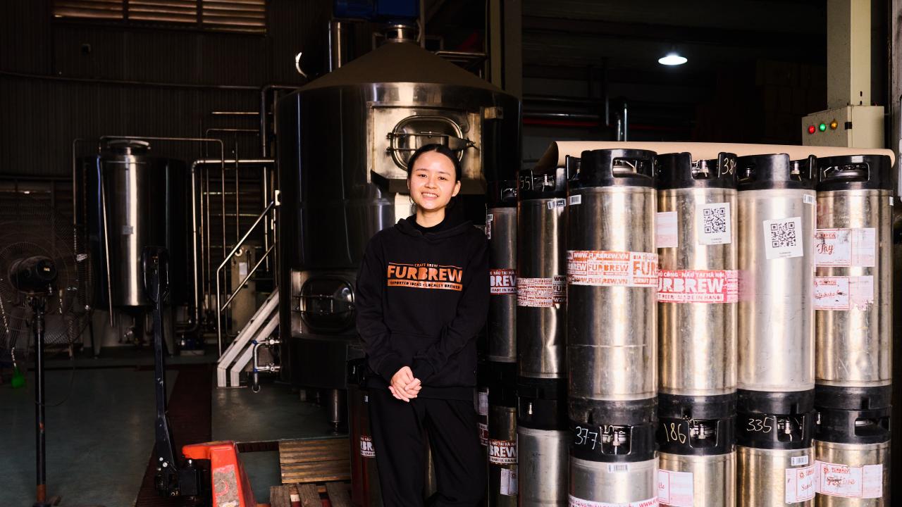 Tapping Potential scholarship winner Dung Ngô stands alongside canisters of beer and other brewing equipment. She wears a black sweatshirt that reads "Furbrew" in orange lettering.