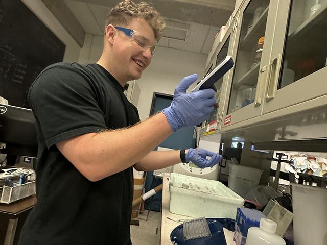 Smiling lab technician wearing gloves and goggles pipettes into a microtube at a bench