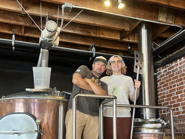 Two brewers embracing beside copper brewing kettles in an industrial brick brewery.