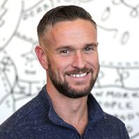 Headshot photo of smiling bearded man in dark shirt with sketched whiteboard background