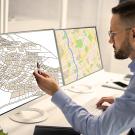 A man analyzes two computer monitors displaying different map layouts in an office setting.