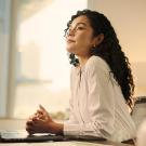 A woman with curly hair sits pensively by a laptop, bathed in soft sunlight.